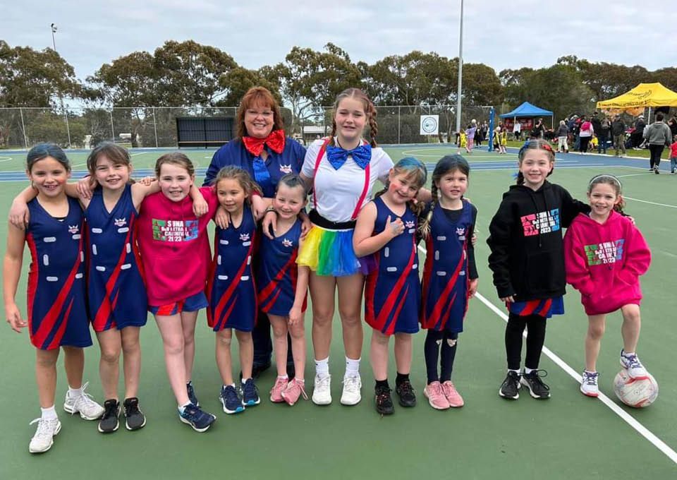 Youth netball team posing on an outdoor court, wearing blue-and-red uniforms with a coach behind them.