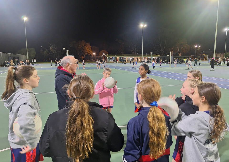 Team gathered on a lit sports field at night, listening to a coach in a blue jumper.
