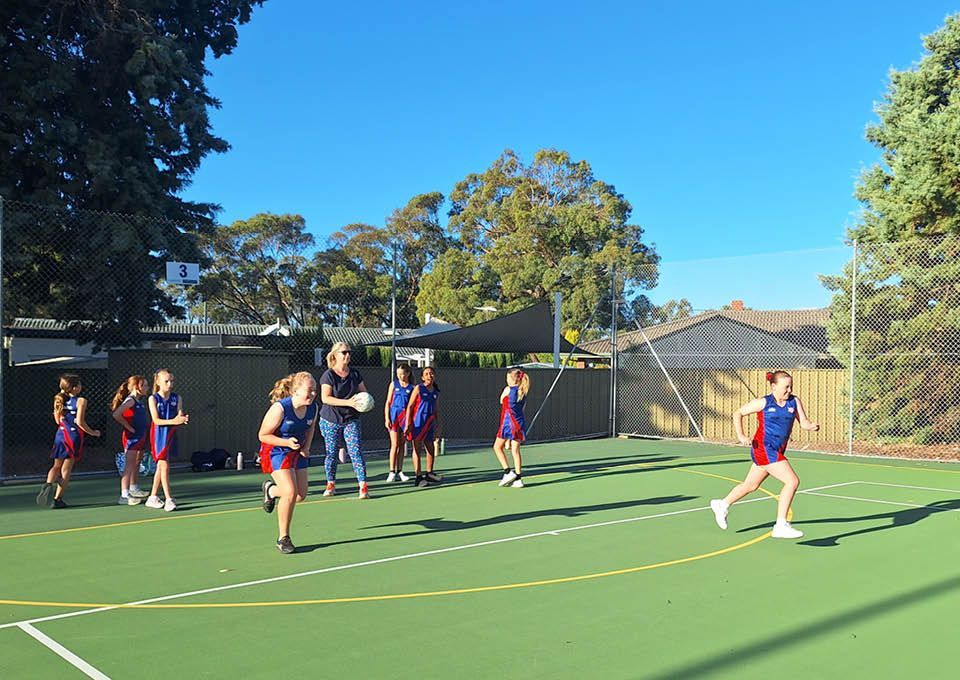 Girls performing netball drills with coach on an outdoor court, with players in blue and red uniforms near the goal.