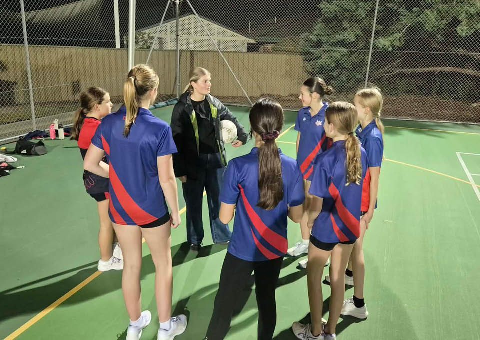 A coach talks to a group of athletes in blue and red uniforms on an outdoor court at night.