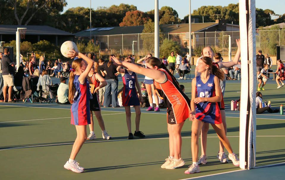 Netball players in red and blue contest a ball near the goal post on an outdoor court.