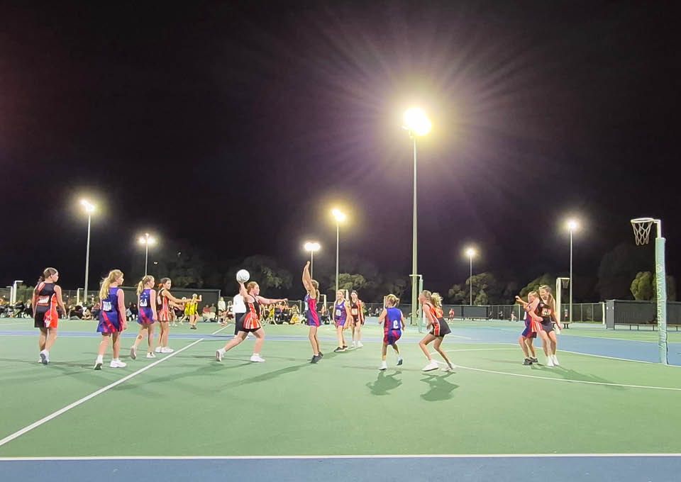 Nighttime netball game on a lit outdoor court, with players in colorful uniforms near the goal.