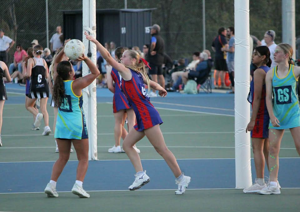 Players contest a ball under the net during an outdoor netball game on a court.