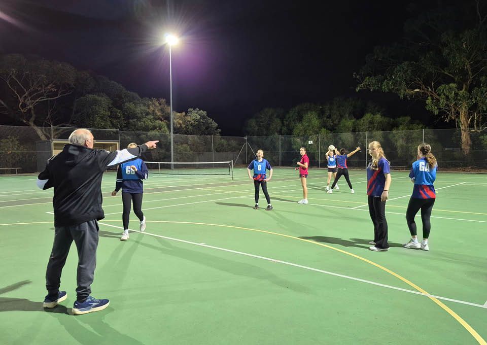Nighttime outdoor netball practice on a lit court with several players and a coach.
