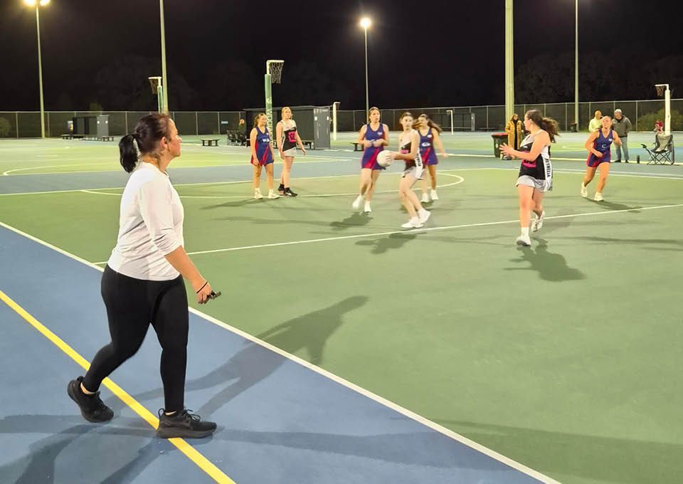 Outdoor night time netball game with players in blue and orange uniforms on a lit court with umpire observing