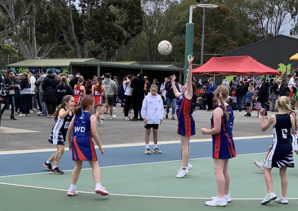 People playing netball on an outdoor court, with spectators and a red canopy in the background.