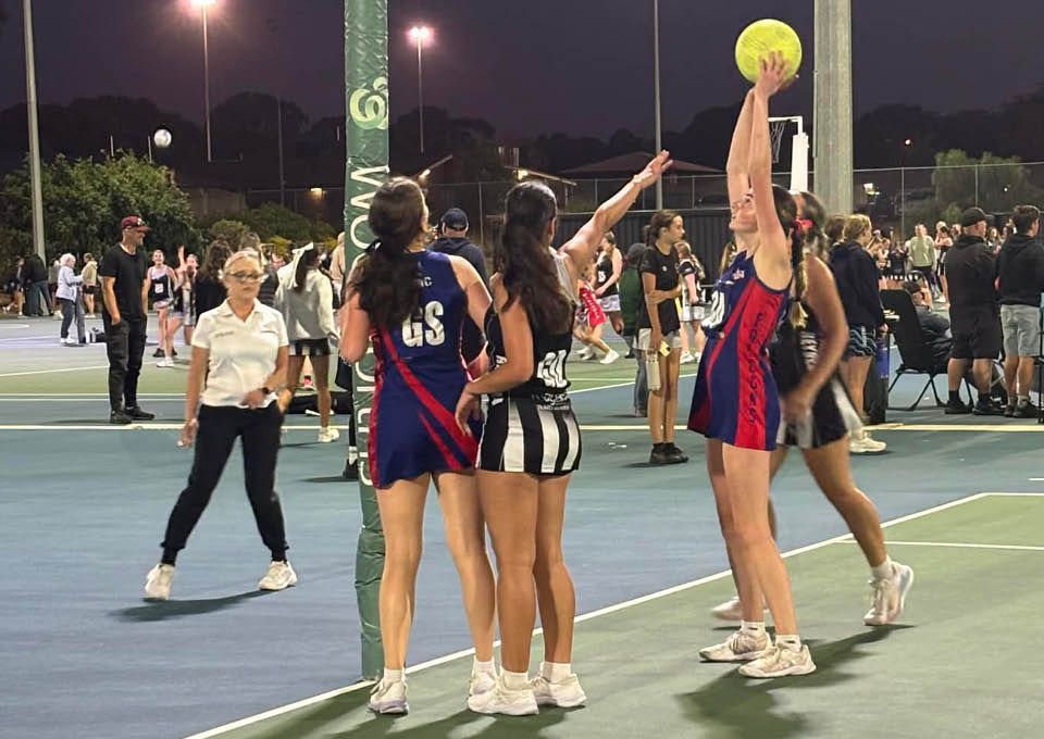 Netball players in red and blue uniforms about to shoot for a goall on an outdoor court at night while an umpire is watching