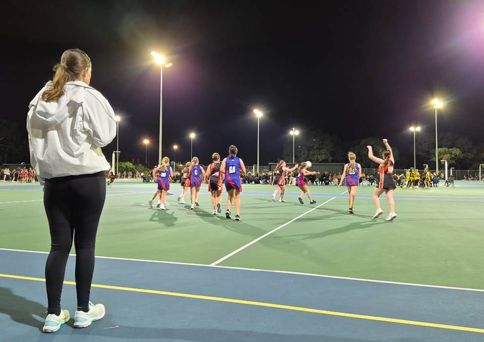 Women playing netball on an outdoor court at night, with a coach watching from the sideline.