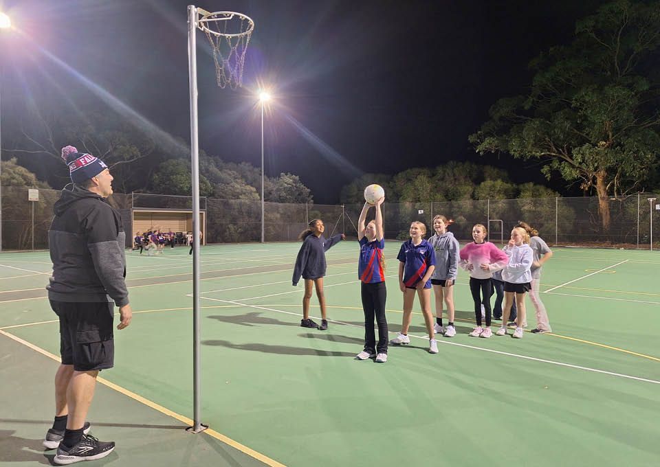 Coach watches players lined up on an outdoor netball court at night.