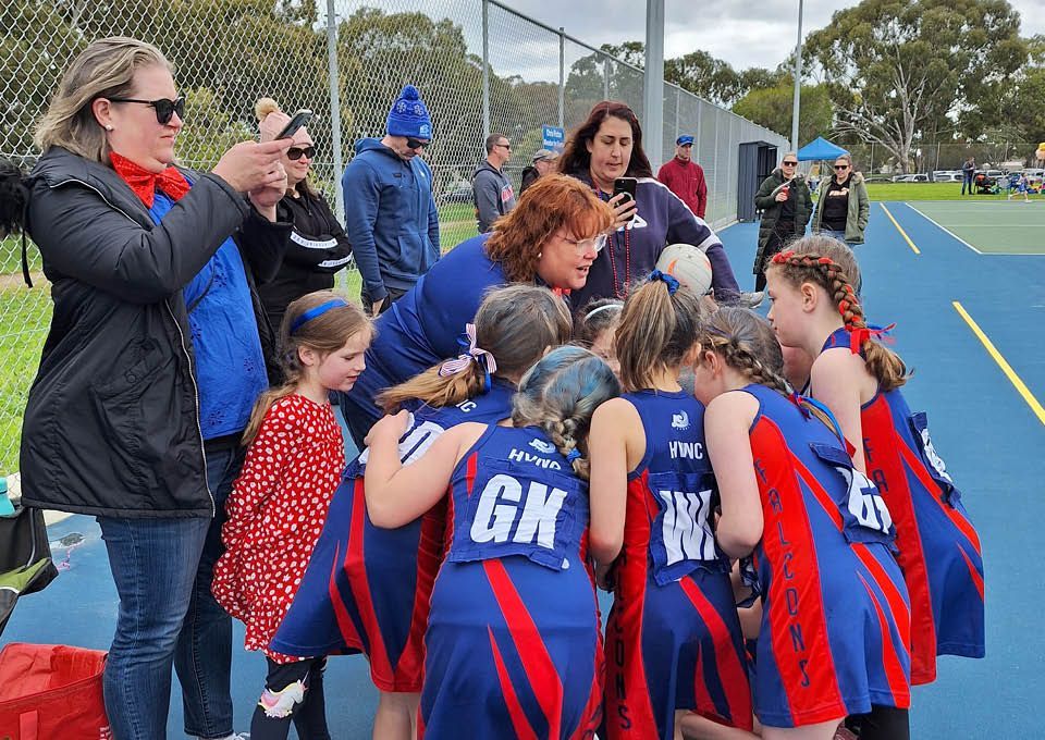 Youth sports team huddle on a blue turf field while spectators watch nearby.