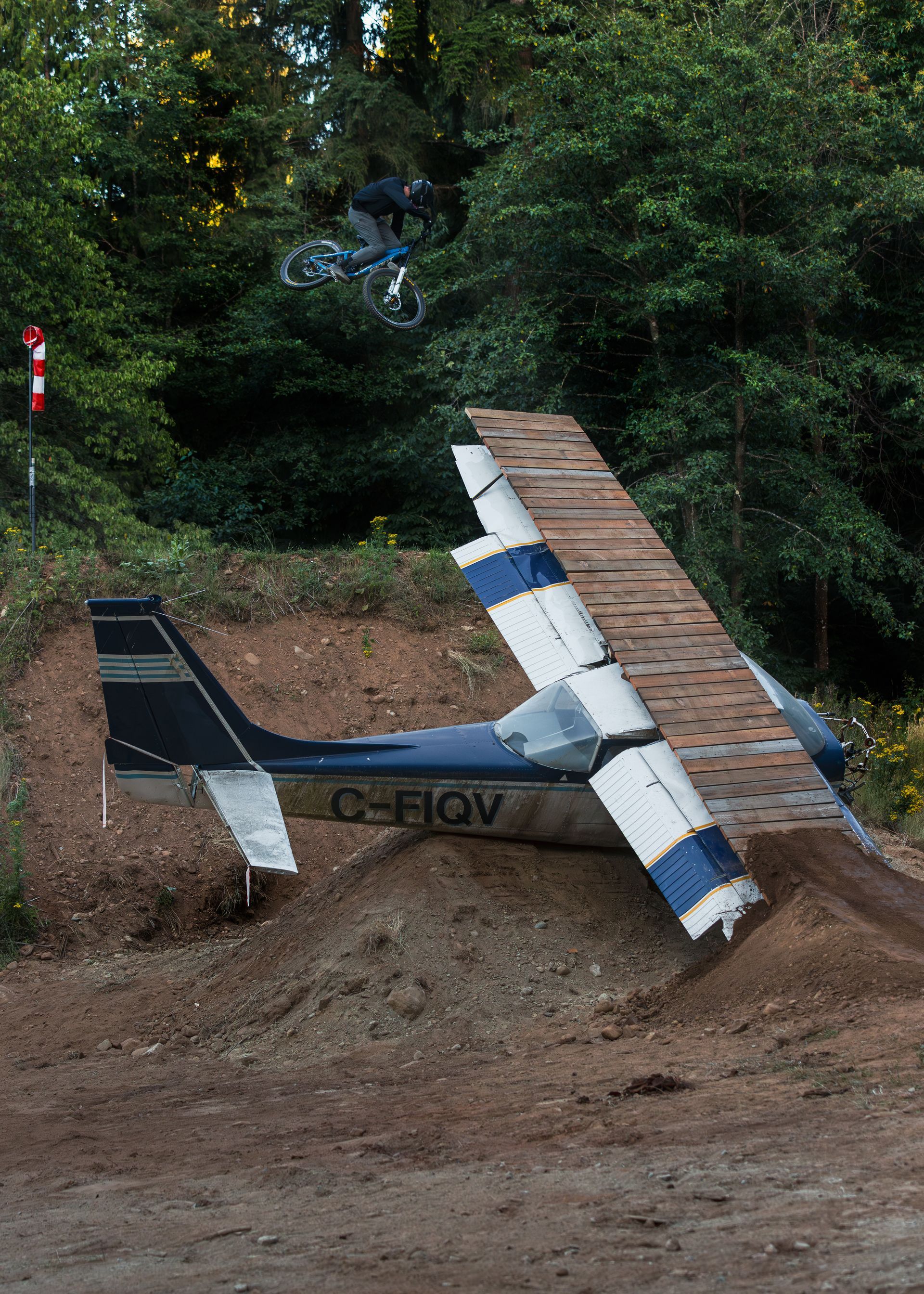 A small plane is wrecked, resting on a dirt hill, wing damaged and leaning against a makeshift wooden structure.