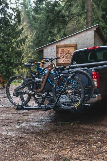 Two mountain bikes mounted on a hitch rack on the back of a truck, parked near a small wooden shed in a forest.