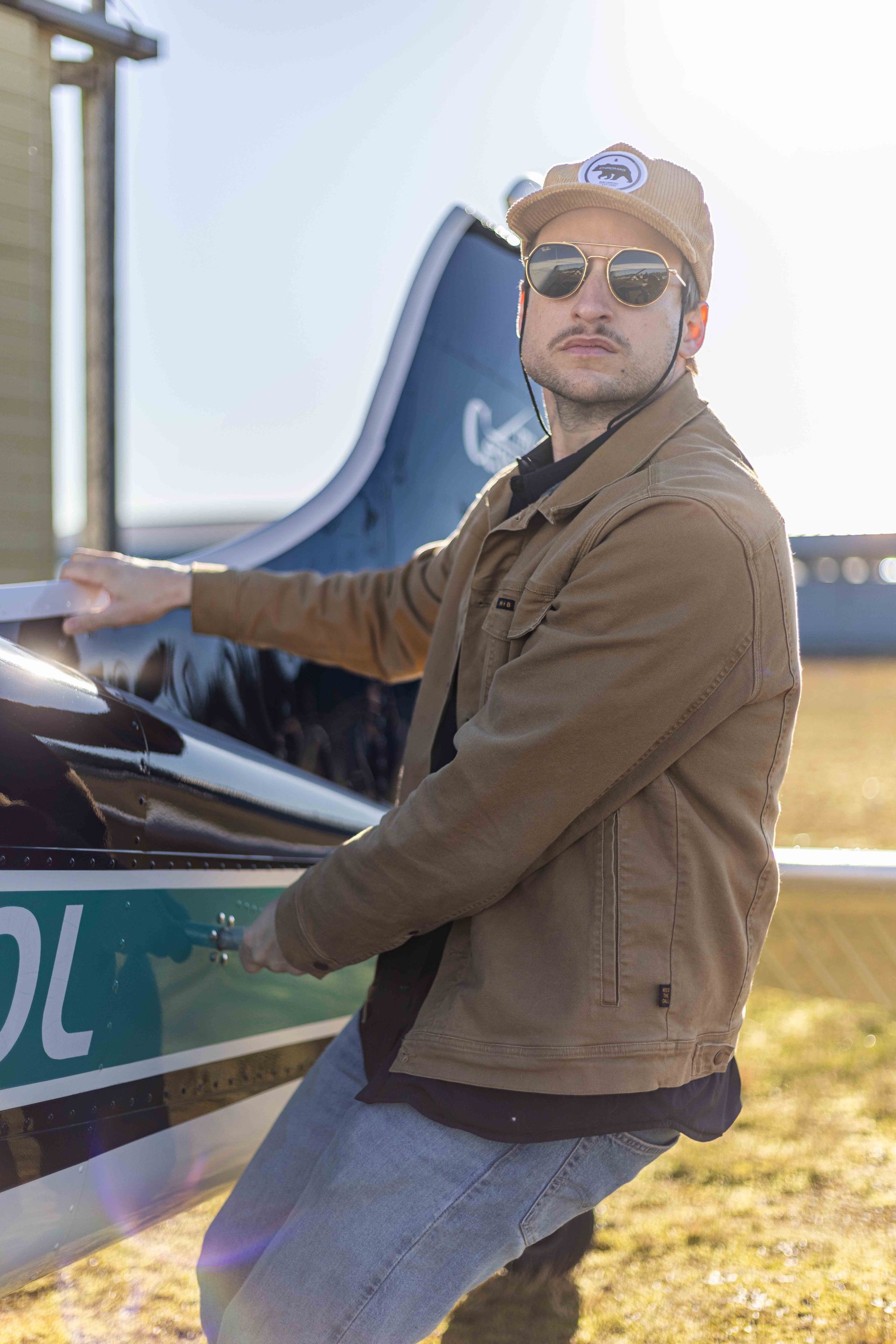 Man in brown jacket and hat, sunglasses, leans against a small airplane, sunny outdoor setting.
