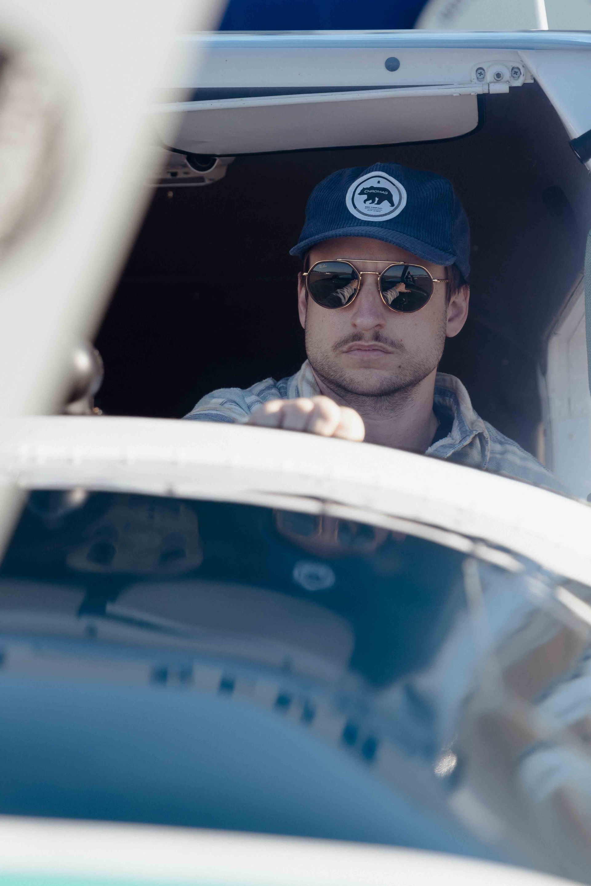 Man wearing sunglasses and a hat in a small plane cockpit, looking forward.