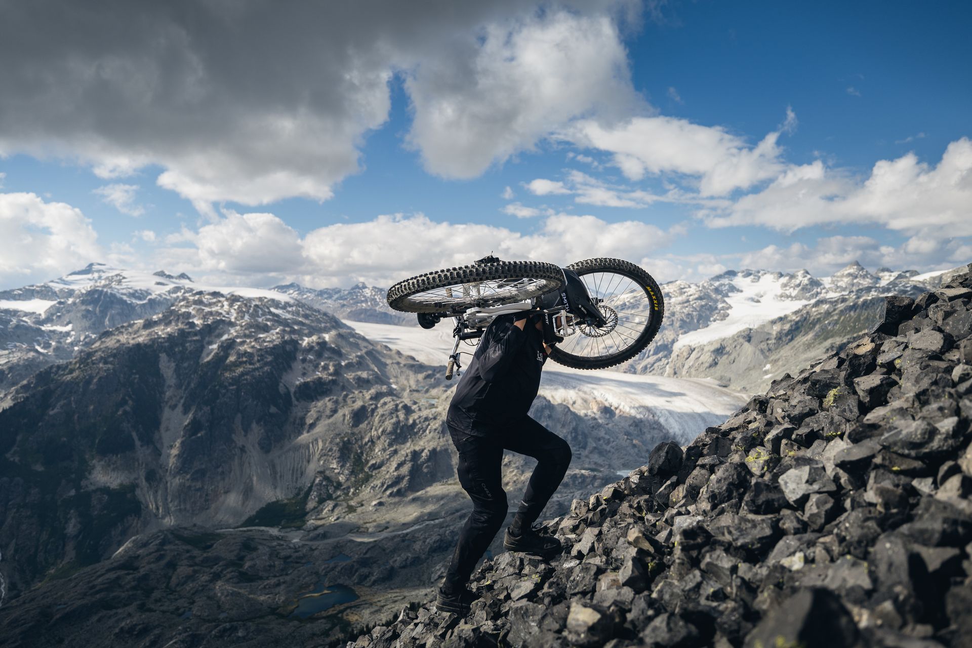 Person carrying mountain bike up a rocky, mountainous slope under a cloudy sky.