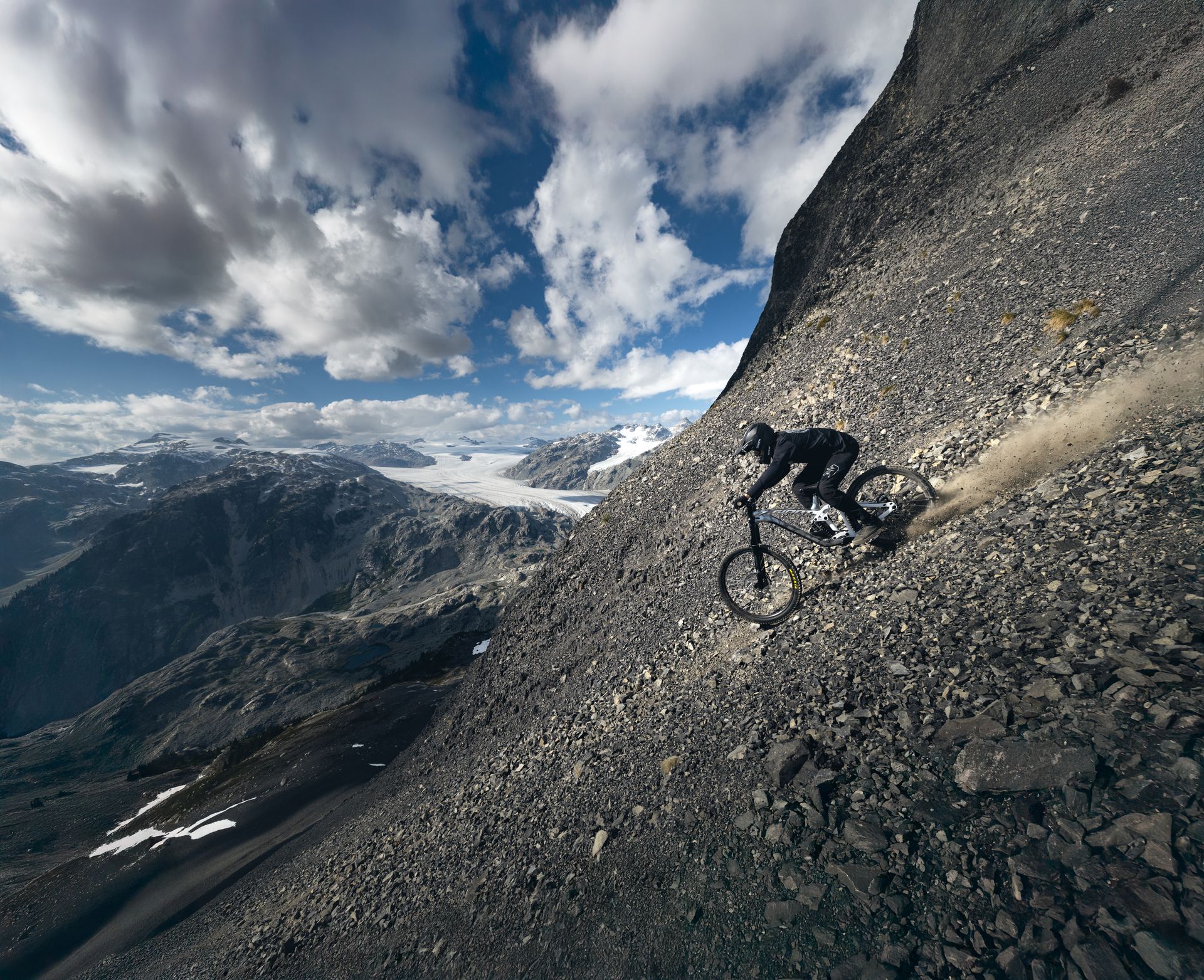 Mountain biker descends a steep, rocky slope with a mountain range and glacier in the background.