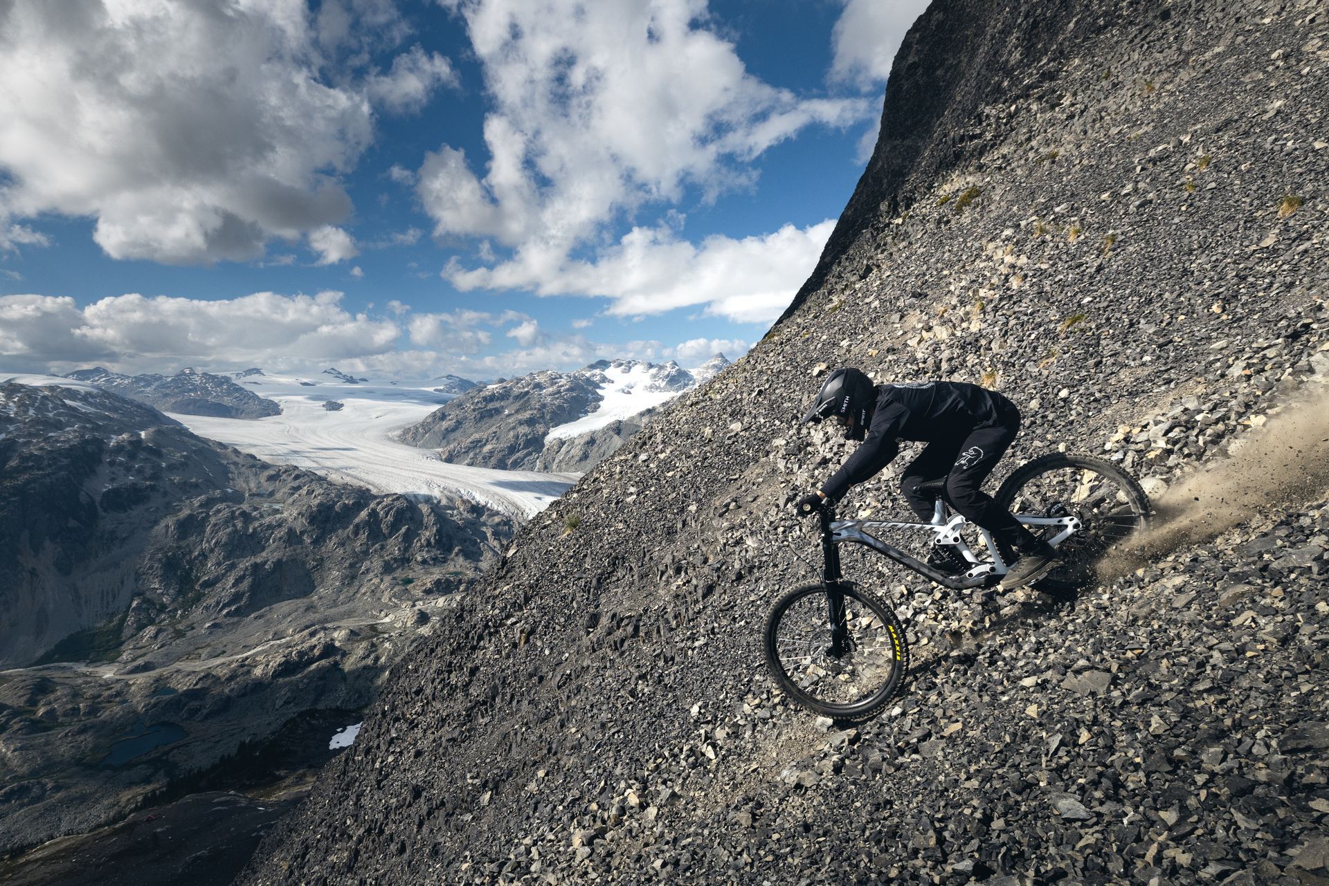 Mountain biker riding down a rocky mountain slope, blue sky, glacier in background.