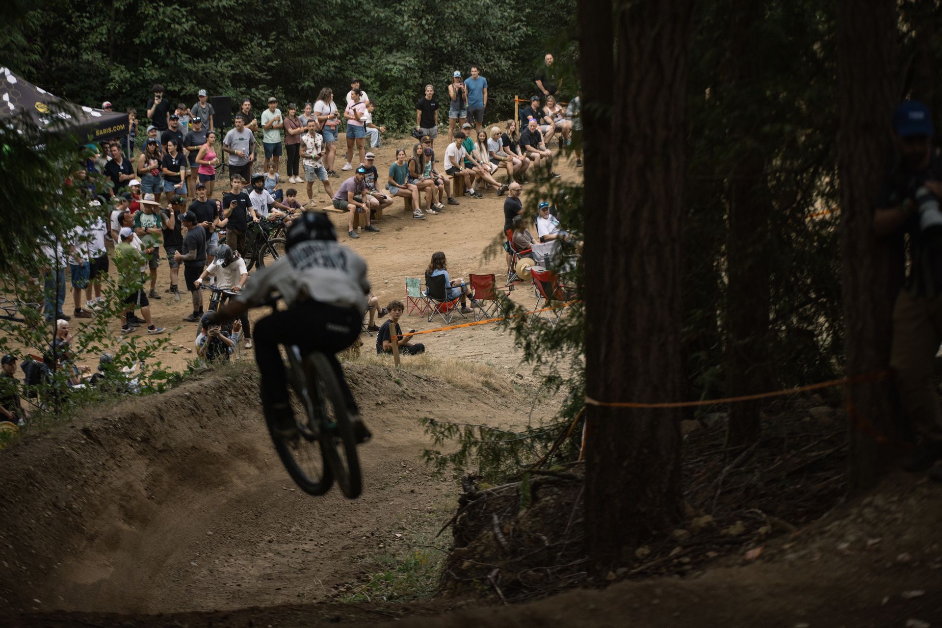 Mountain biker in mid-air jump on dirt trail, trees and crowd in the background.