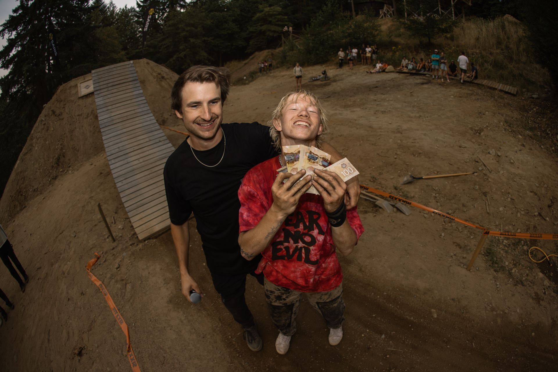 Two men smile, holding money, at a dirt bike jump in an outdoor setting.