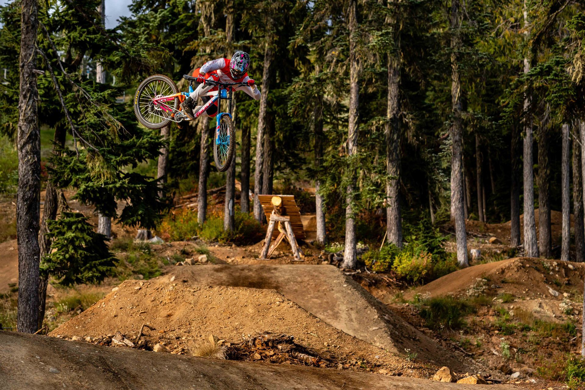Mountain biker in mid-air, jumping over a dirt ramp in a forest setting.