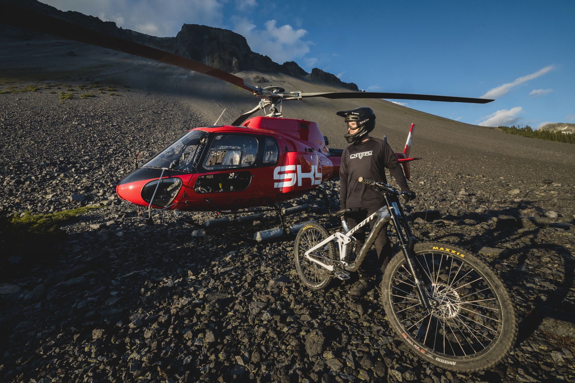 Mountain biker standing next to a red helicopter on a rocky mountain slope.