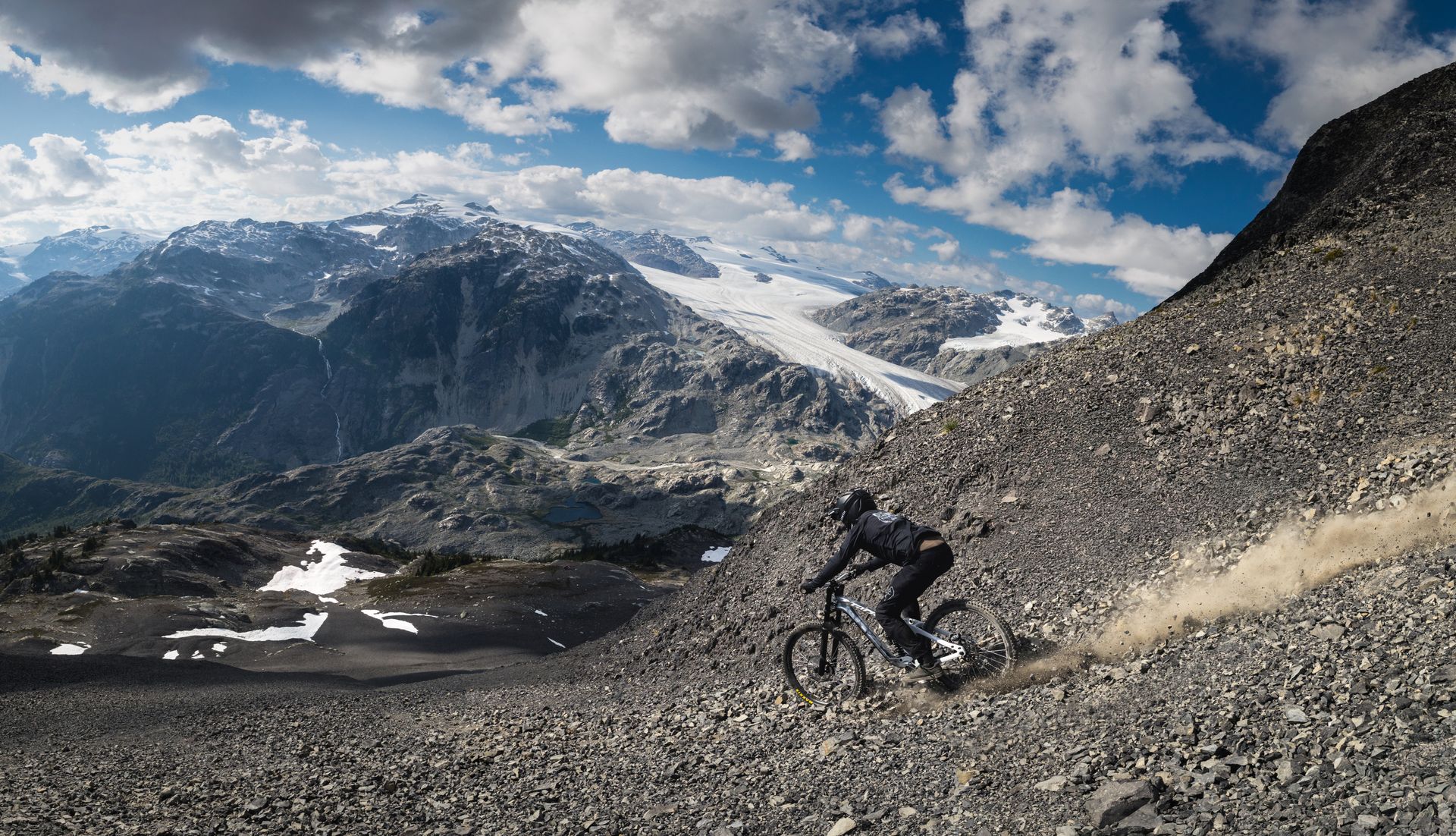 Mountain biker descending a rocky mountain slope, with snow-capped peaks and cloudy sky in the background.