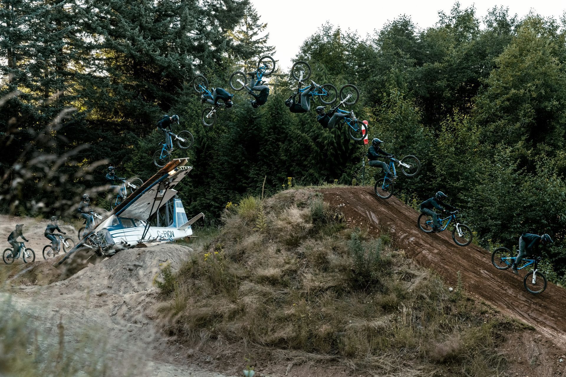 Mountain biker performing aerial tricks over a dirt jump in a wooded area.
