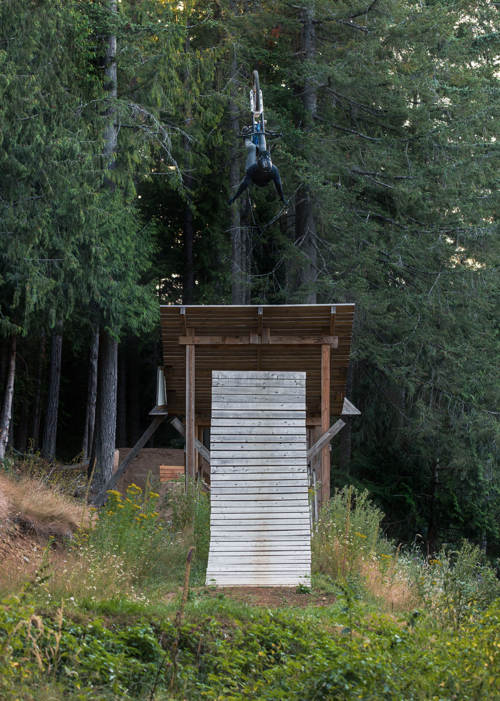 Mountain biker in mid-air, performing trick off a wooden jump in a forest setting.