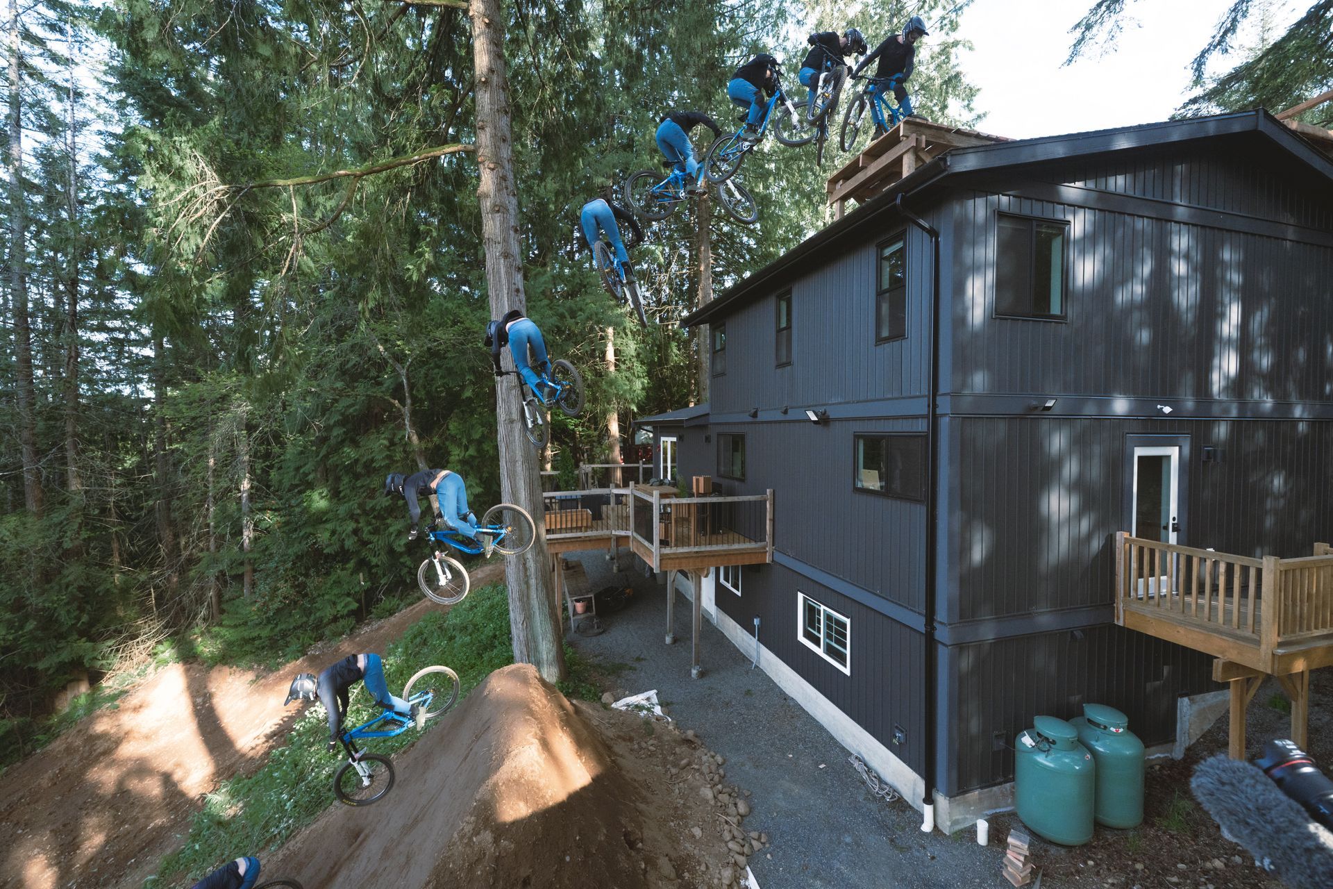 Mountain biker performing multiple jumps, near a dark-colored house in a wooded area.