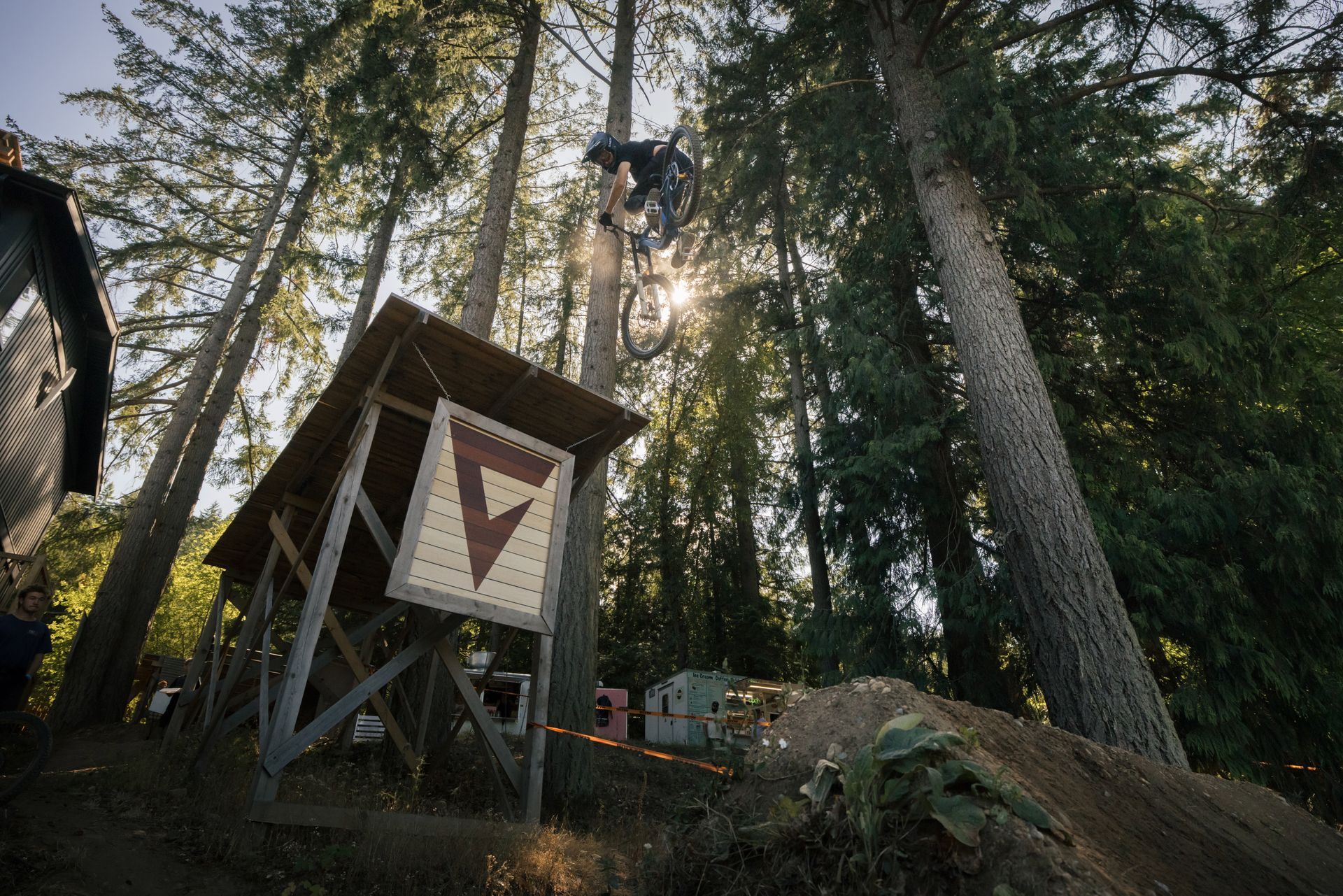 Mountain biker mid-air over wooden ramp in a forest, sun shining through trees.