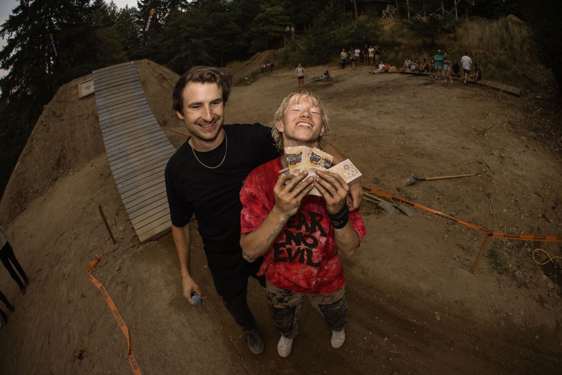 Two men on a dirt hill smiling, one holding money, other's arm around him. Outdoor setting, overcast.