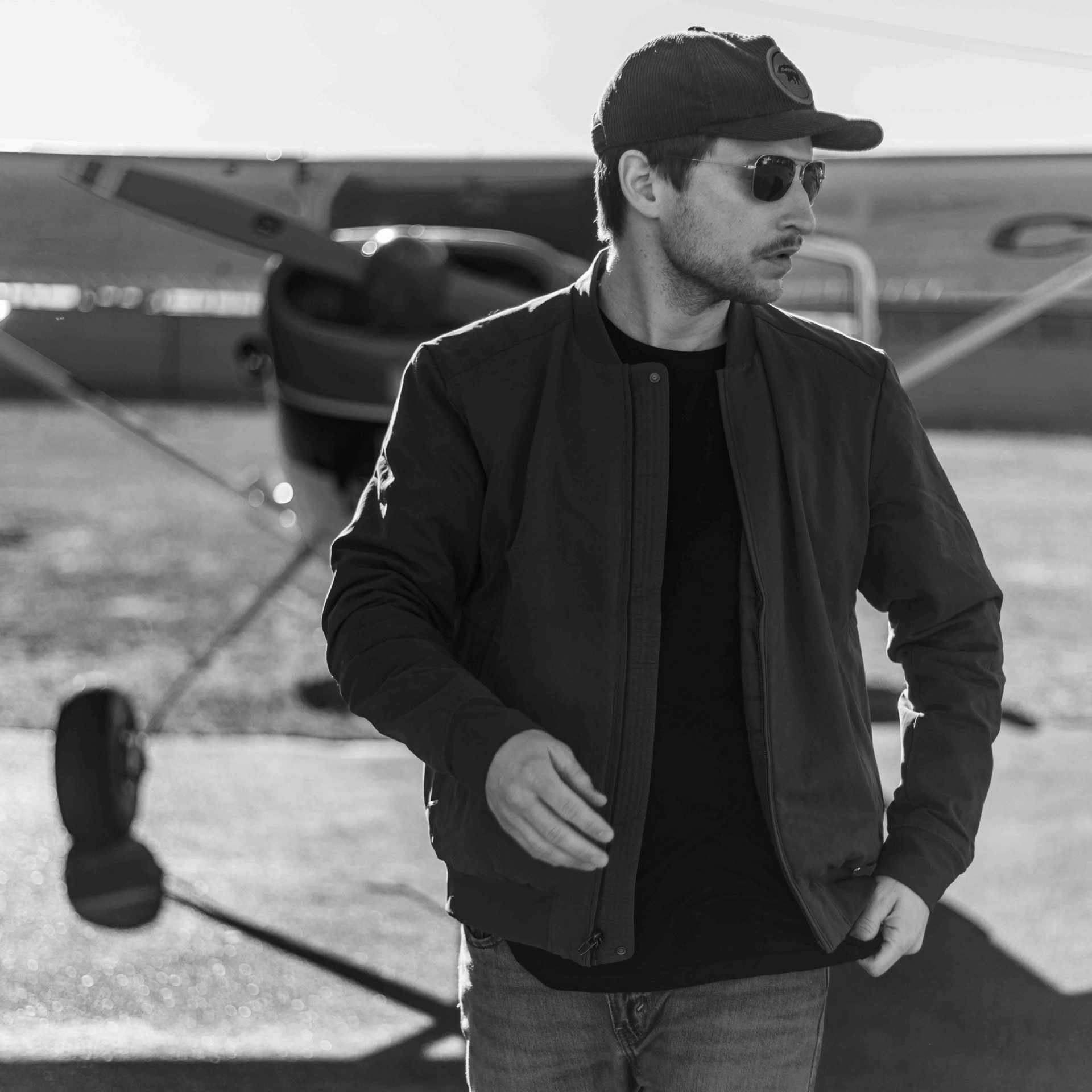 Man in bomber jacket and sunglasses stands near an airplane.