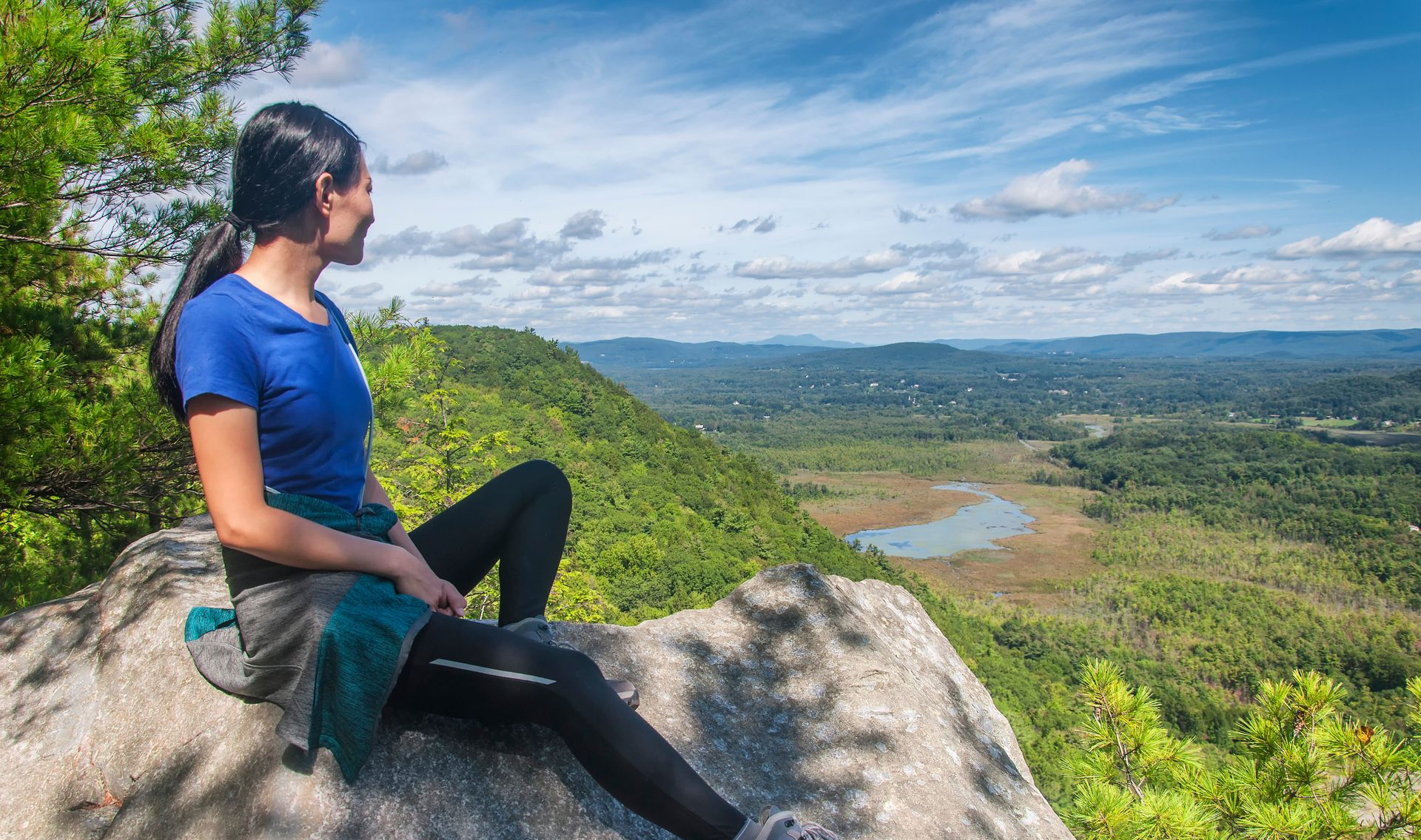 Woman in blue shirt sits on a rock, looking at a scenic valley.