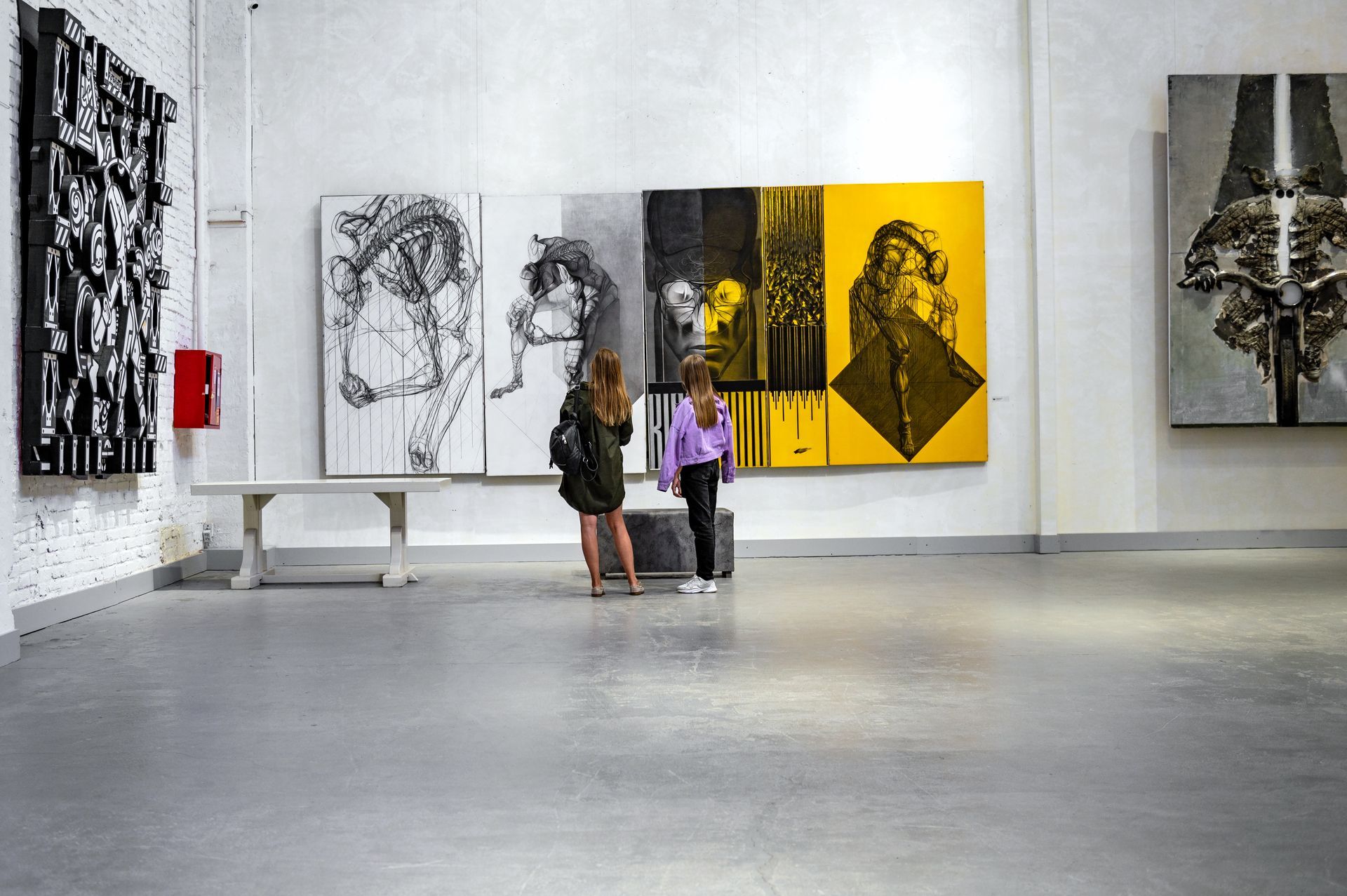 Two people looking at abstract art in a gallery; white walls, gray floor.