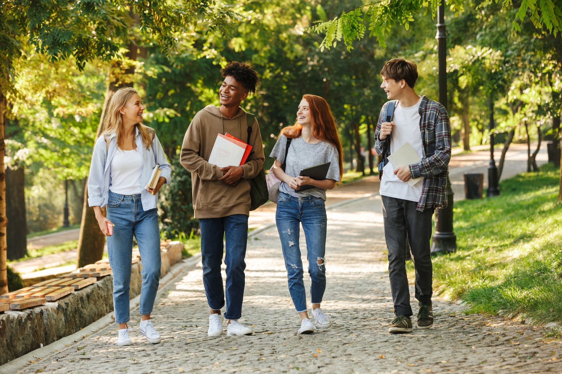 Four students walk along a sunny park path, carrying books and backpacks, laughing and talking together.