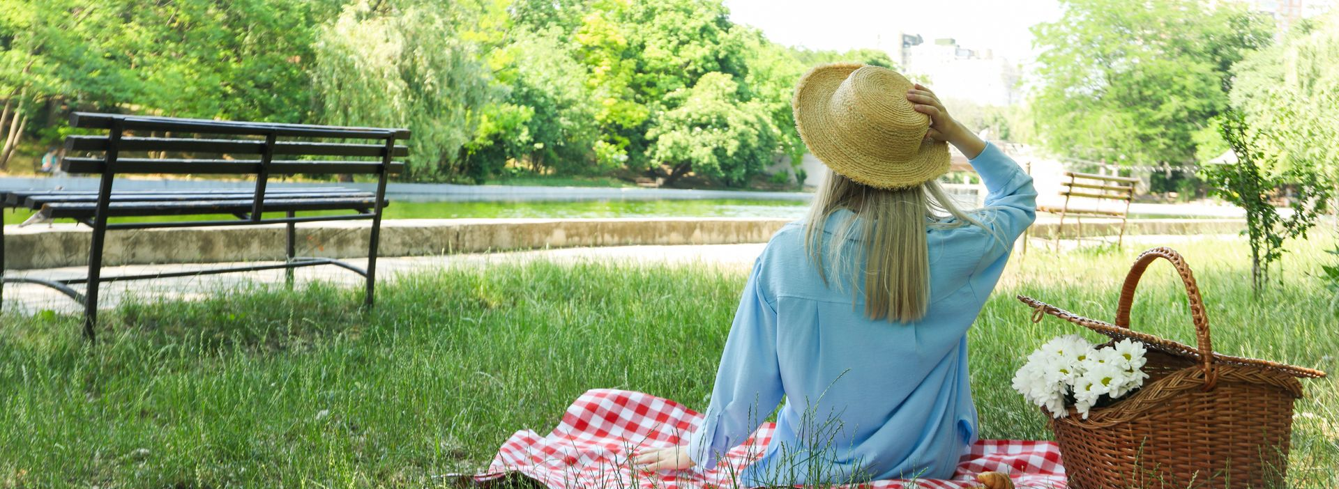 Woman sitting on a picnic blanket in a sunny park, facing a lake with a bench nearby.