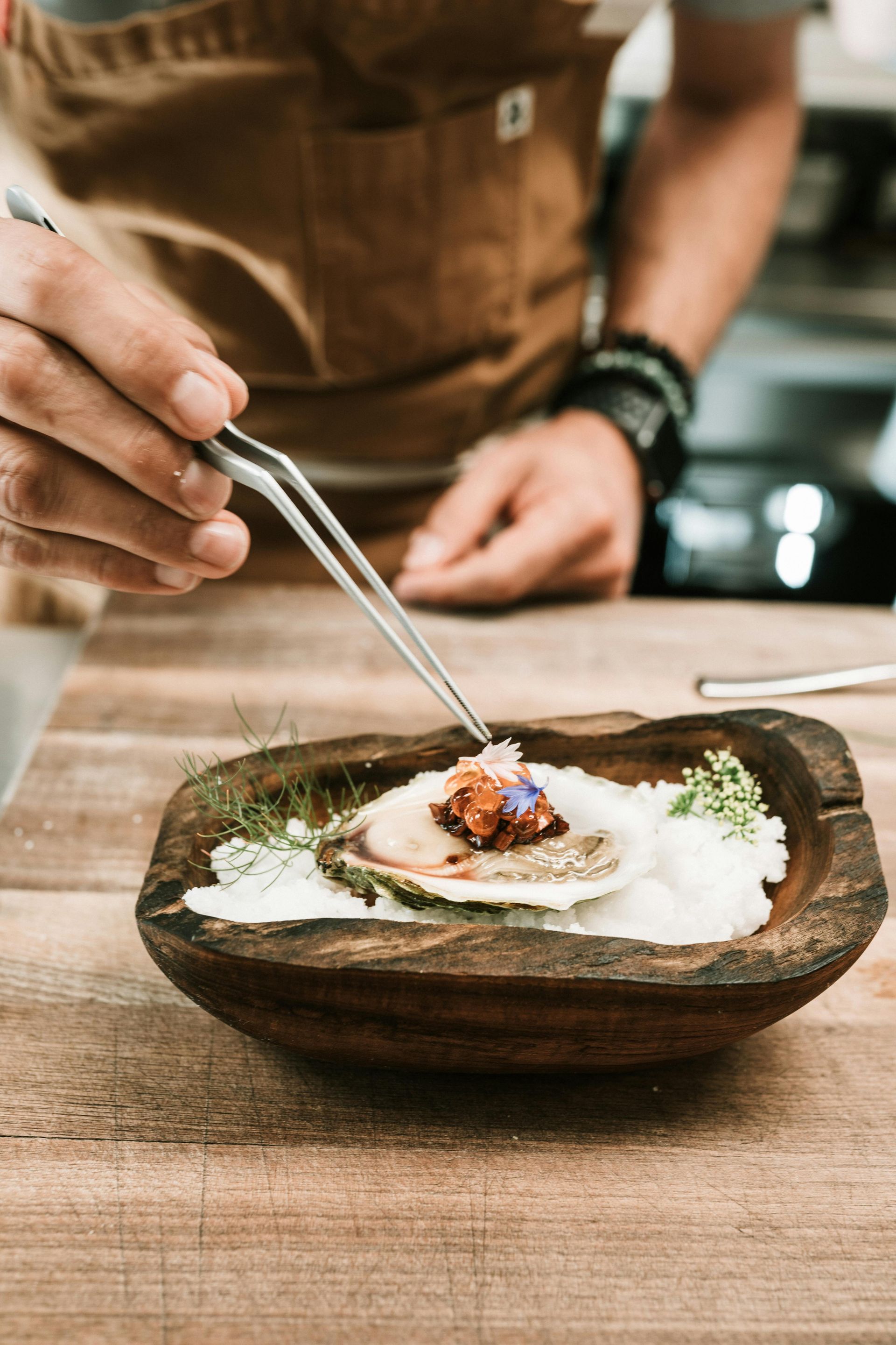 A person is preparing food in a wooden bowl with tweezers.
