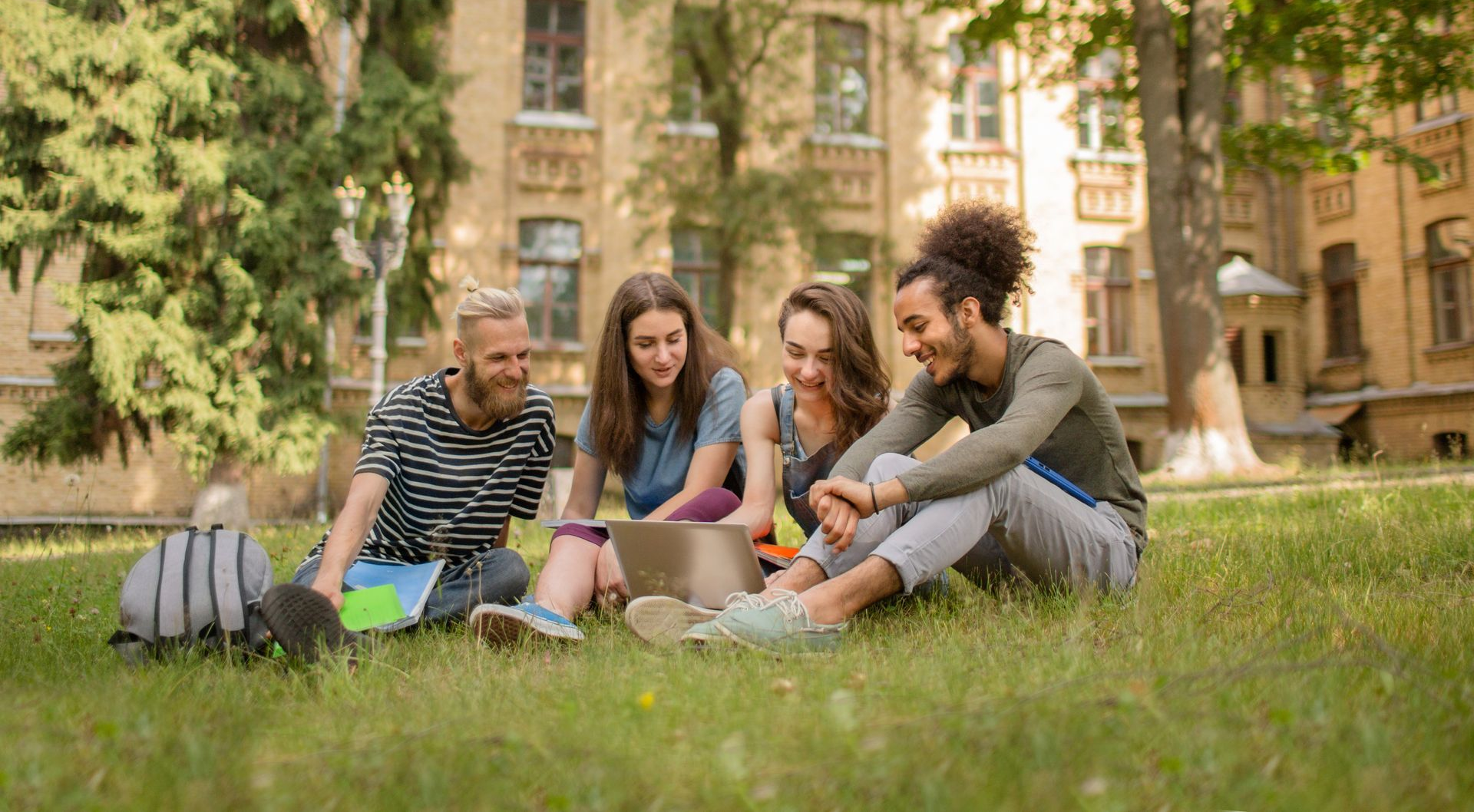 Four students sit on grass outside a university building, looking at a laptop together and smiling.