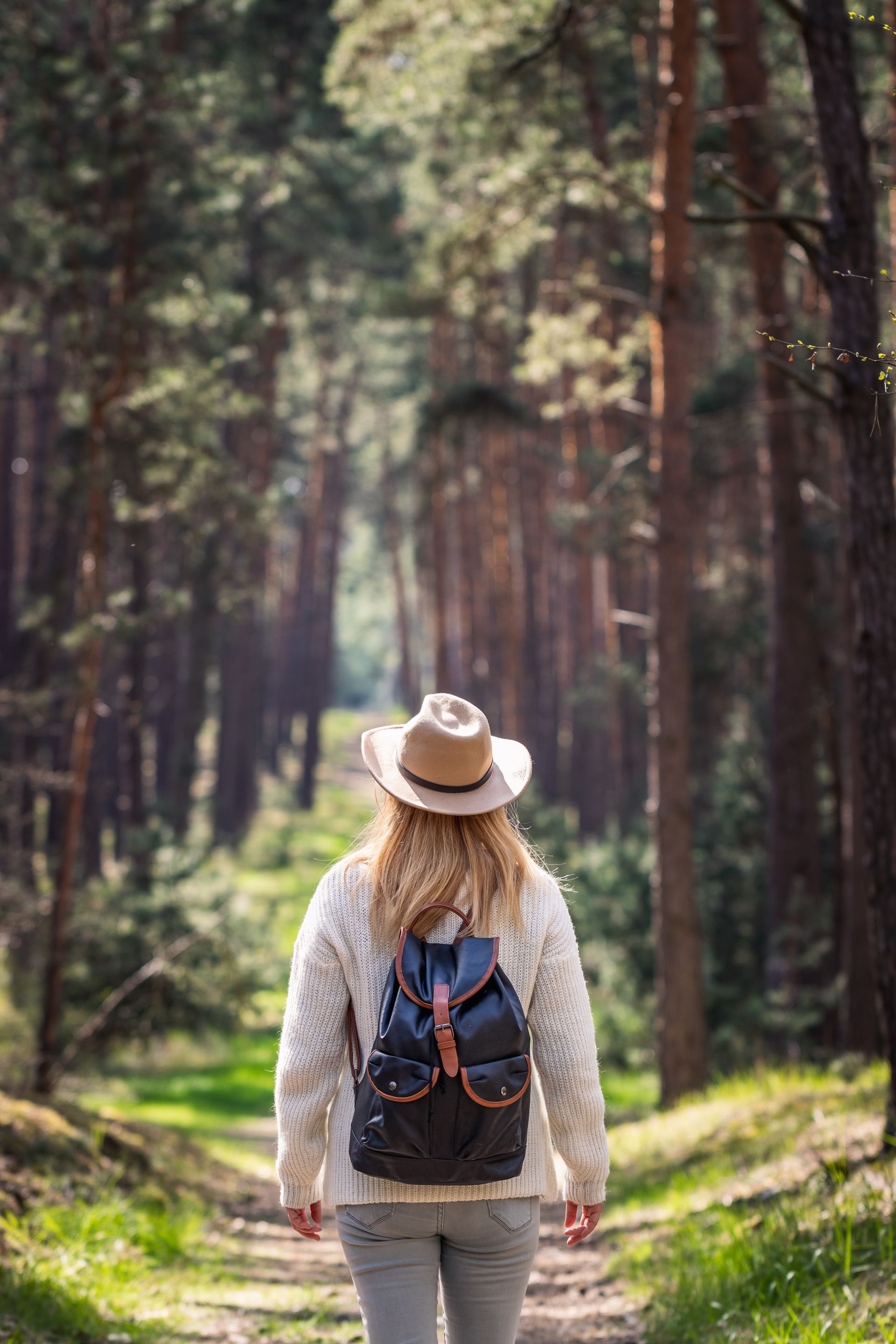 Person hiking on a forest trail with a backpack, wearing a hat, surrounded by tall trees