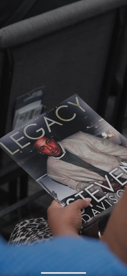 Hand holding a black book titled “LEGACY,” featuring a man in a light suit on the cover.