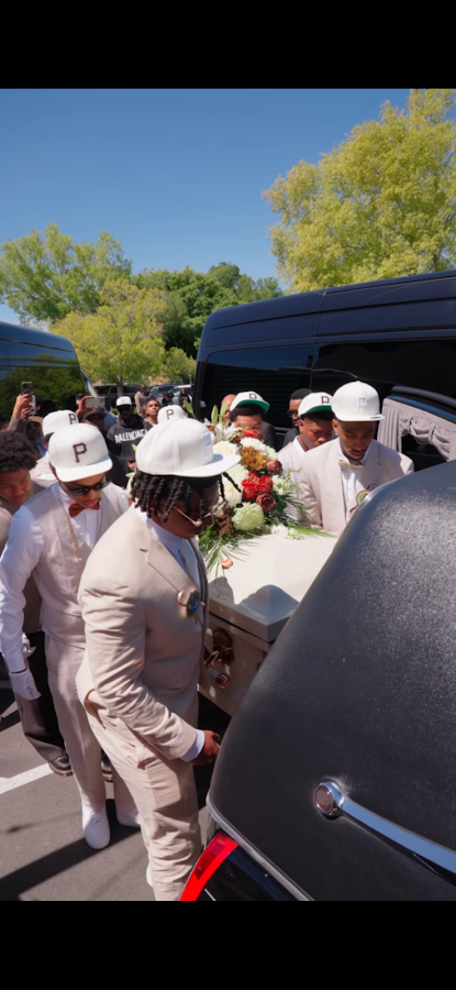 People in white uniforms and hats gathered beside a black vehicle in bright sunlight, carrying flowers.