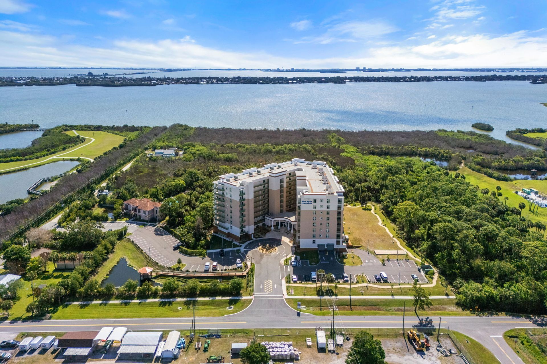 Aerial view of a waterfront condominium complex beside a road and green parkland.