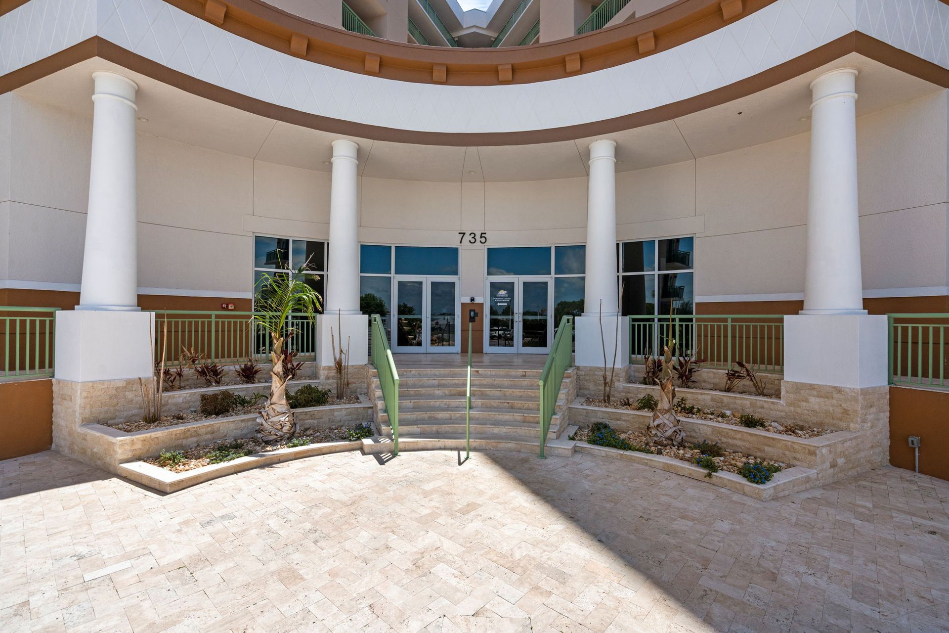 Front entrance of a modern building with white columns, glass doors, and steps leading up from a landscaped courtyard