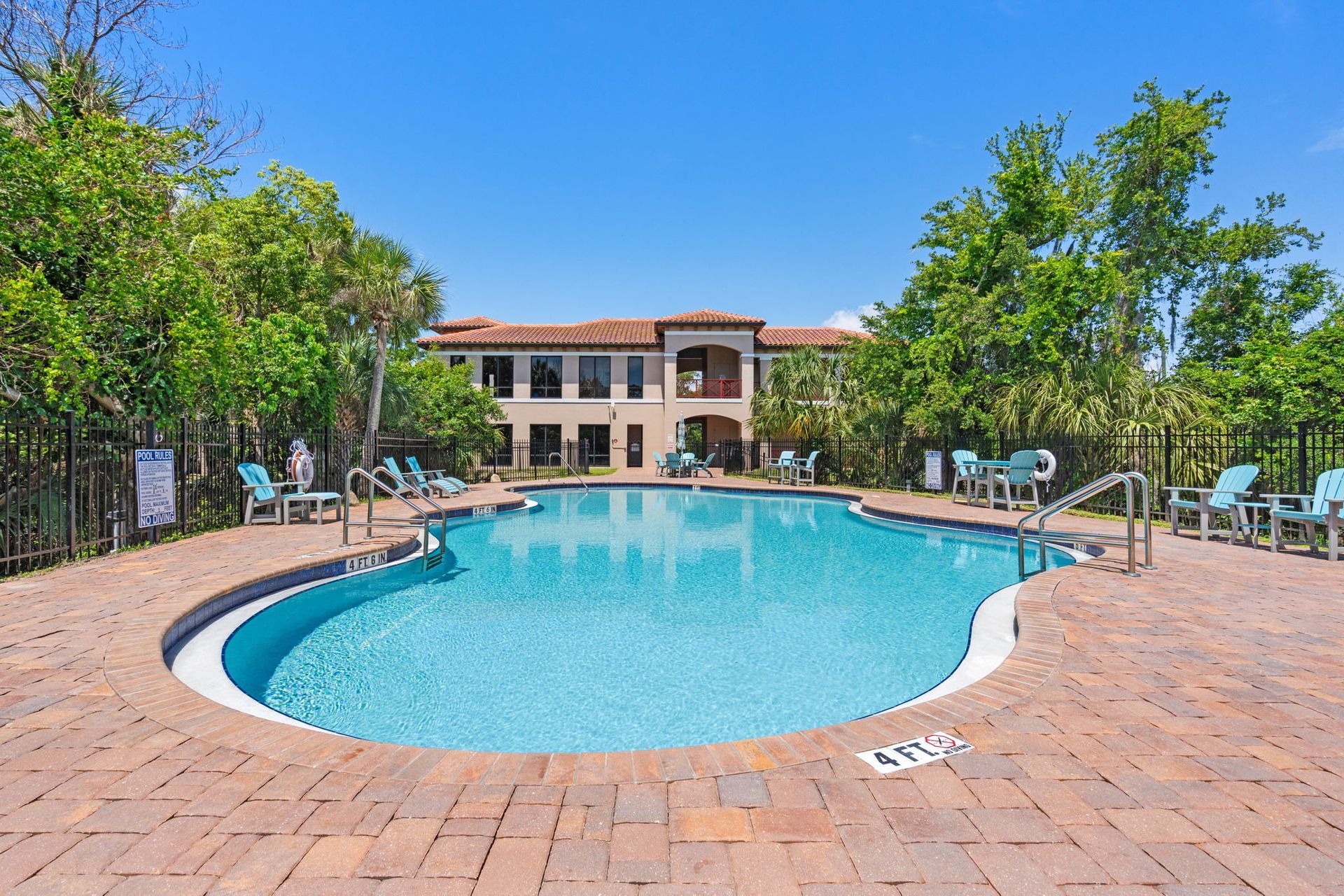 Outdoor resort pool surrounded by brick patio and lounge chairs, with a hotel building and trees in the background