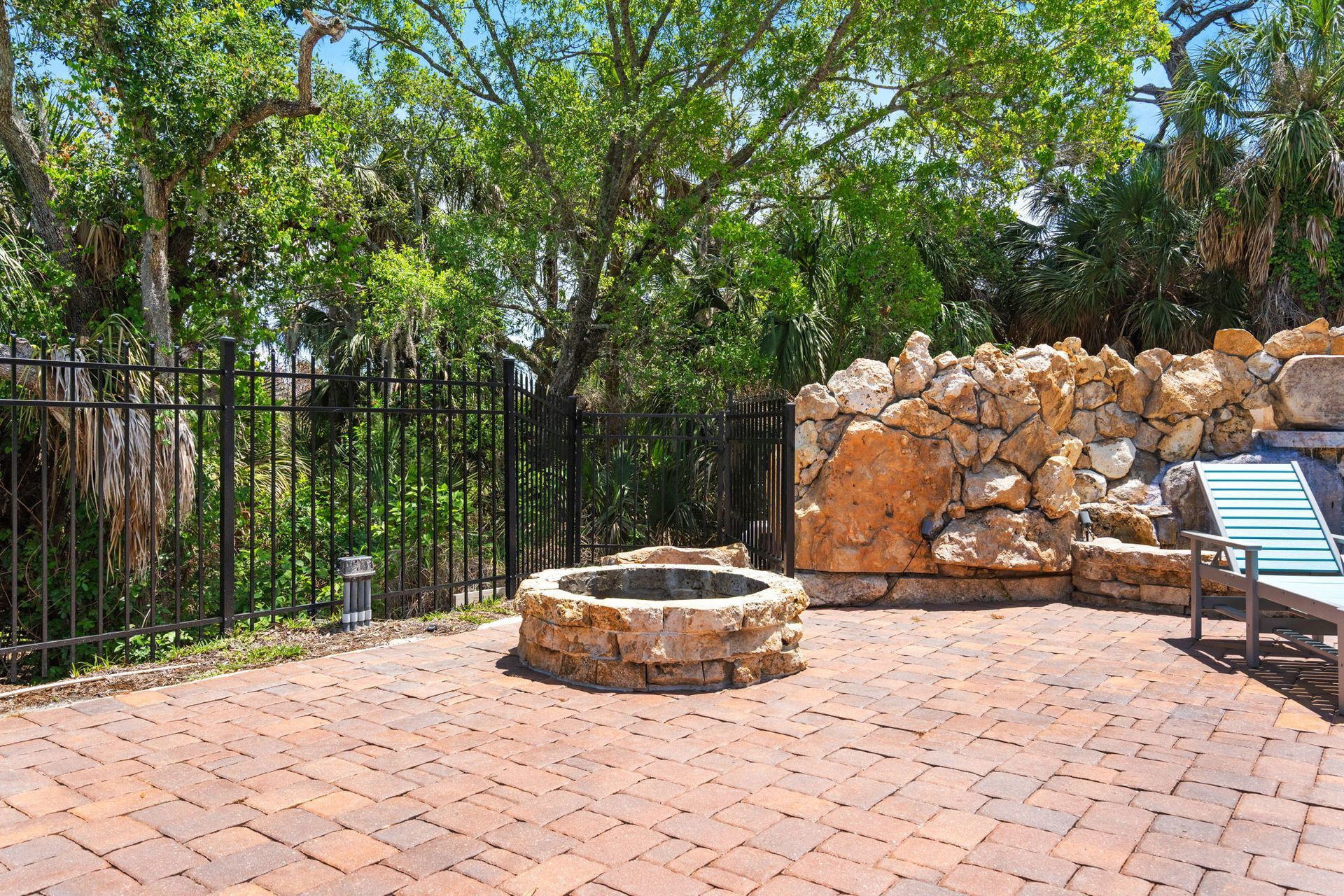 Patio with stone fire pit, black fence, rock wall, and lounge chair in a sunny garden area
