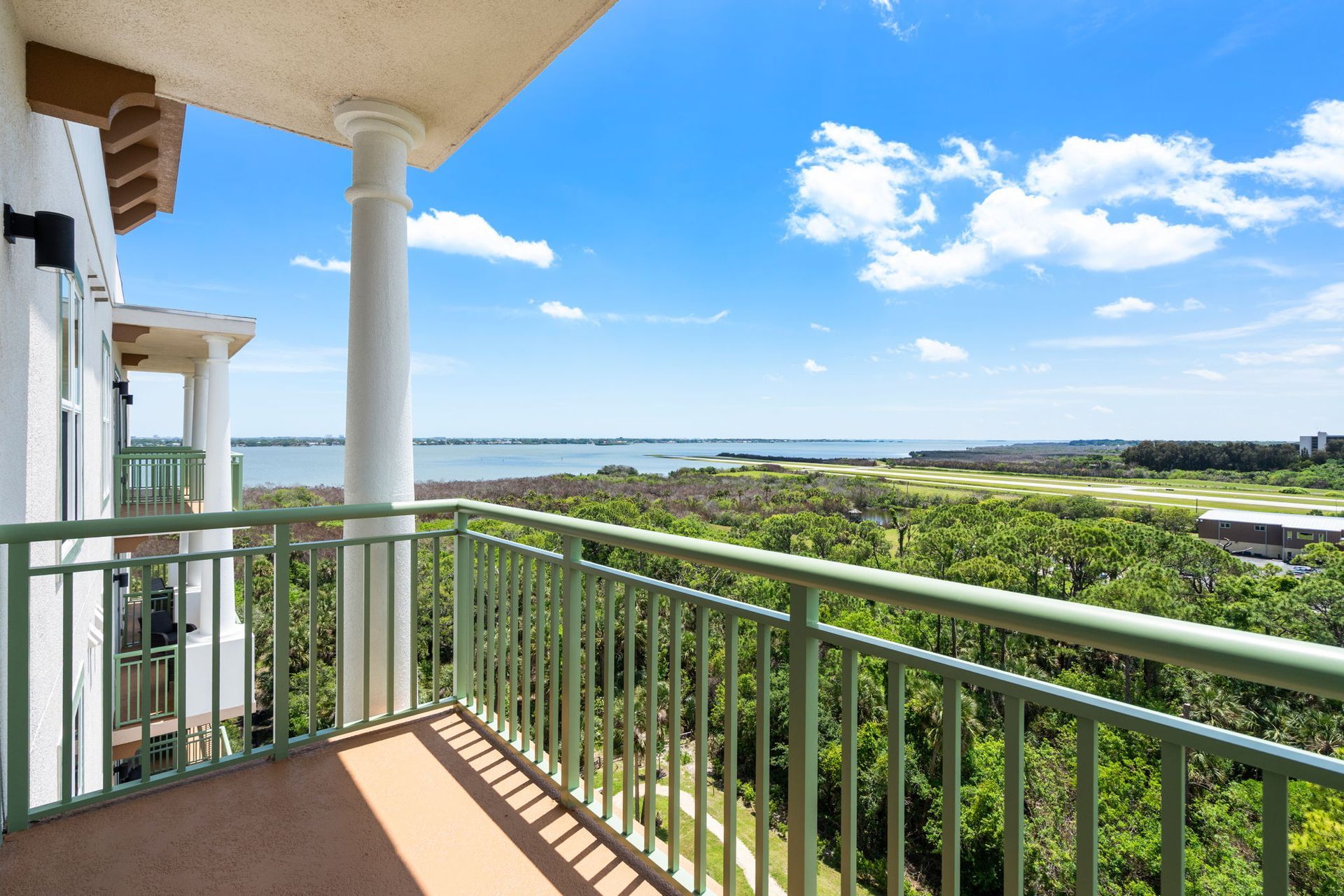 Balcony with green railing overlooking lush trees, water, and a bright blue sky with clouds