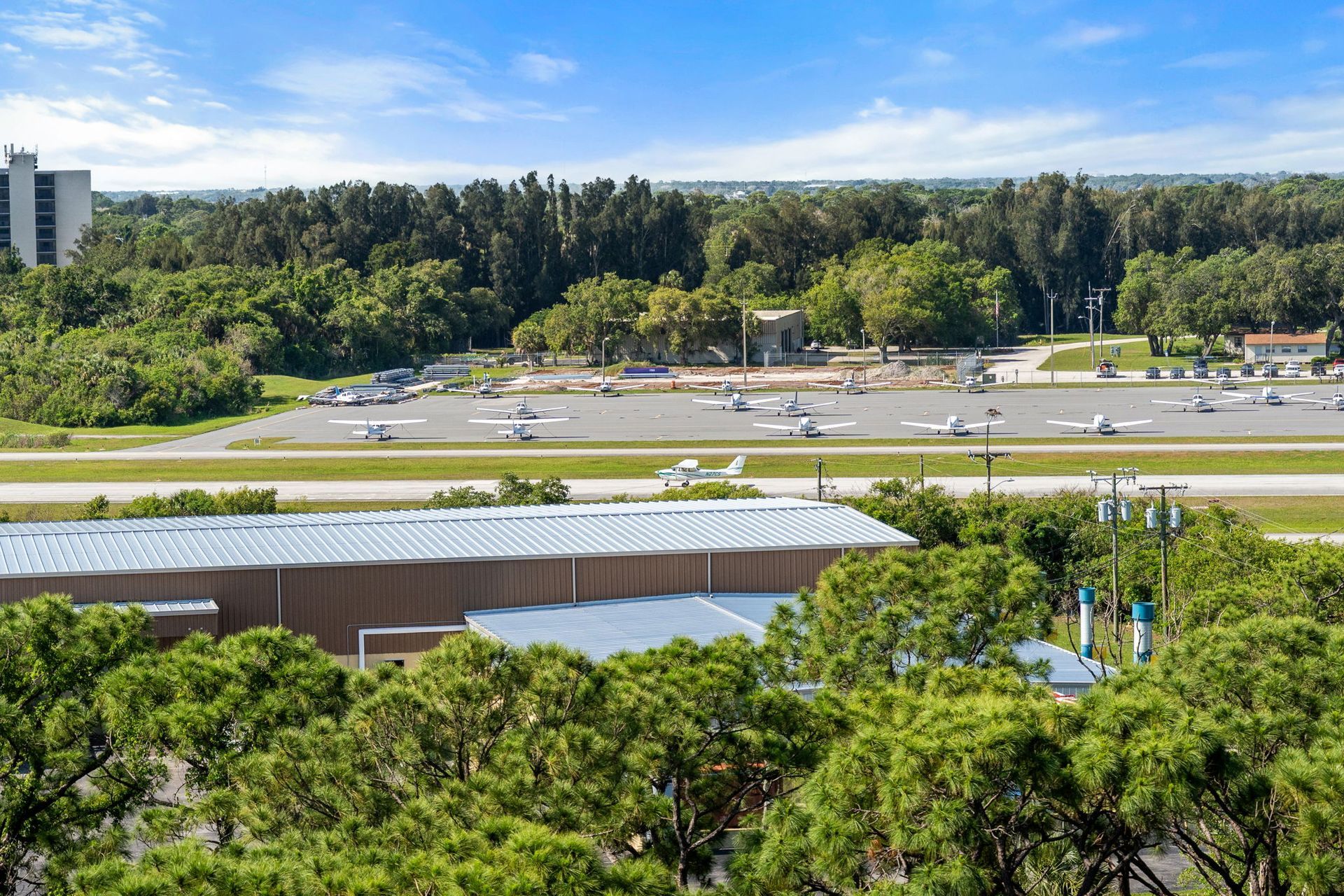 Aerial view of a parking lot beside a road, with dense green trees and a building in the foreground.