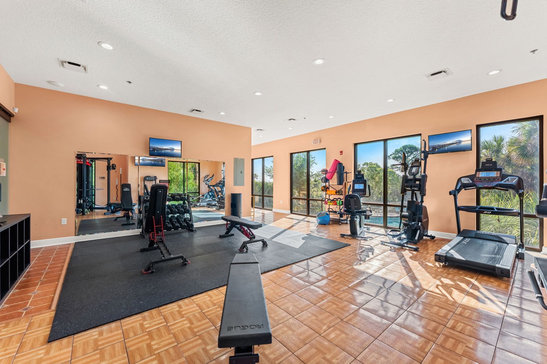 Bright gym with exercise machines, mirrors, windows, and tiled floor