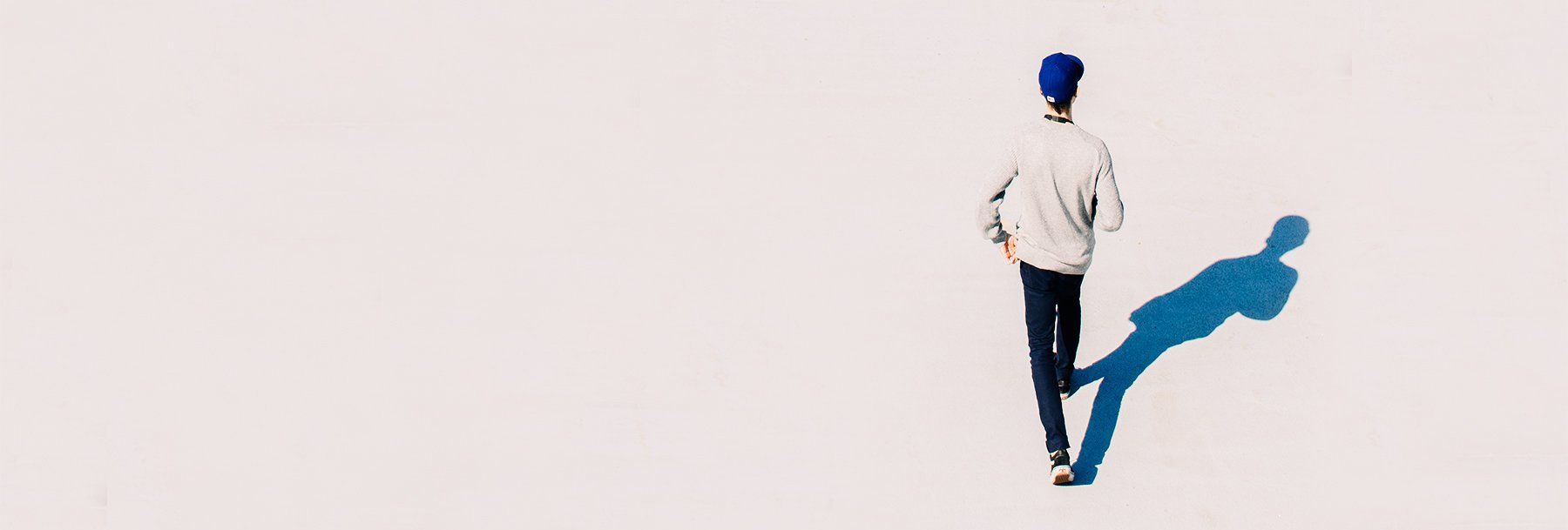 A man in a blue hat is walking with his shadow on the ground.