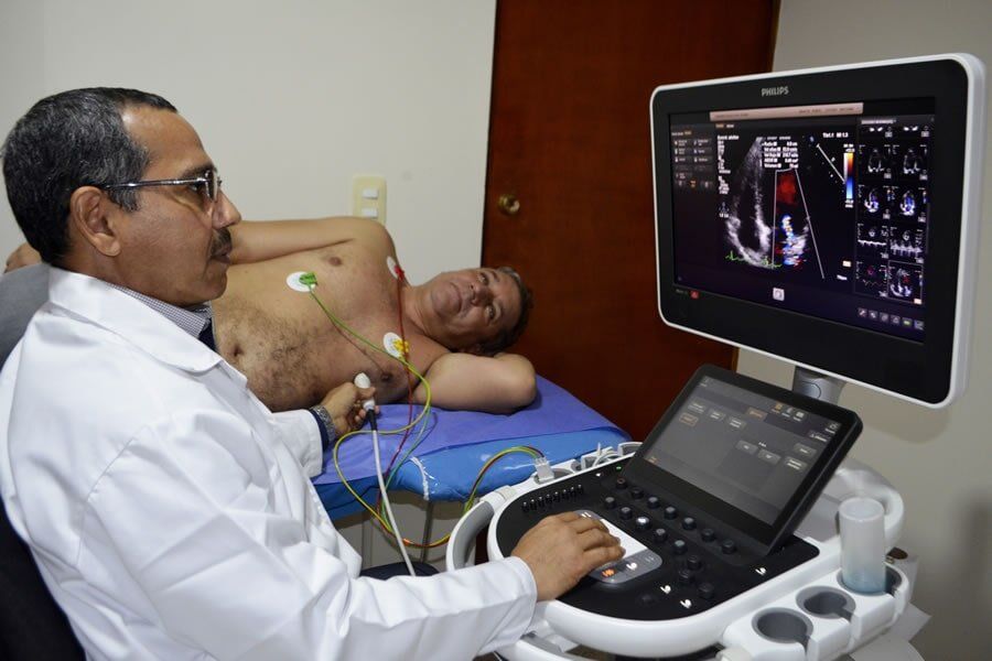 A medical professional performs an echocardiogram on a patient lying on an exam table in a clinical setting.